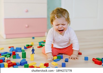 Upset Crying Baby Girl With Educational Toys. Sad Tired Or Hungry Alone Healthy Child Sitting Near Colorful Different Wooden Blocks At Home Or Nursery. Baby Missing Mother In Daycare