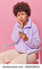 Upset Curly Haired Woman Sits On Chair Uses Mobile Phone Received Bad Sms Feels Very Unhappy Wears Fur Jacket And Trousers Isolated Over Pink Background Feels Offended After Quarrel With Boyfriend