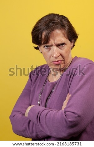 Upset defensive mature woman in purple blouse on a yellow background looking directly at the camera with arms folded scowling.