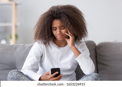 Upset depressed african american teen adolescent girl looking at phone sit on sofa at home, sad teenager cry holding cellphone waiting for call feel bad anxious being rejected scared of cyberbullying