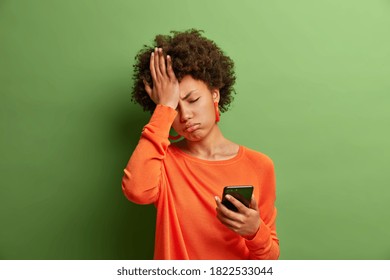 Upset Depressed Afro American Woman Makes Face Palm As Forgot About Something Important Holds Mobile Phone Expresses Sorrow And Regret Blames Herself Isolated On Green Background. Negative Emotions