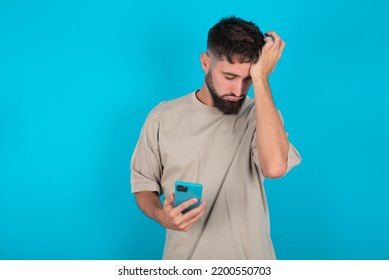 Upset Depressed Bearded Caucasian Man Wearing Casual T-shirt Over Blue Background Makes Face Palm As Forgot About Something Important Holds Mobile Phone Expresses Sorrow And Regret Blames