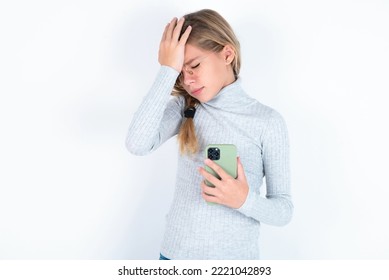 Upset Depressed Beautiful Caucasian Blond Teen Girl Wearing Gray Turtleneck Sweater Over White Background  Makes Face Palm As Forgot About Something Important Holds Mobile Phone Expresses Sorrow 
