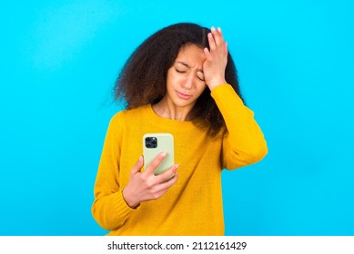 Upset Depressed Beautiful Teen Girl Wearing Yellow Sweater Standing Against Blue Background Makes Face Palm As Forgot About Something Important Holds Mobile Phone Expresses Sorrow And Regret Blames