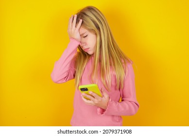 Upset Depressed Caucasian Little Kid Girl Wearing Long Sleeve Shirt Over Yellow Background Makes Face Palm As Forgot About Something Important Holds Mobile Phone Expresses Sorrow And Regret Blames