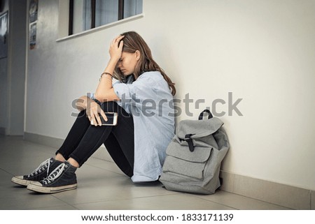 Upset and depressed girl holding smartphone sitting on college campus floor holding head. University sad student suffering from depression sitting on floor at high school. Lonely bullied teen.