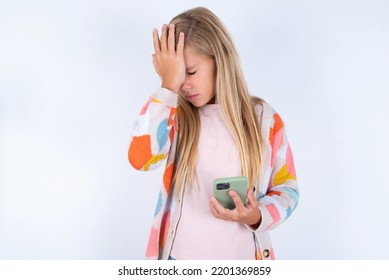 Upset Depressed Little Kid Girl Wearing Colorful Yarn Jacket Over White Background Makes Face Palm As Forgot About Something Important Holds Mobile Phone Expresses Sorrow And Regret Blames