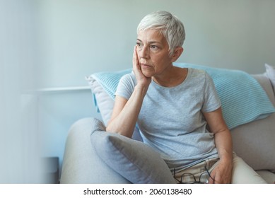 Upset depressed senior gray-haired lady touching temple, sitting on sofa, looking in the window, feeling worried and stressed about the future and thinking about own problems - Shutterstock ID 2060138480