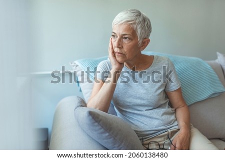 Upset depressed senior gray-haired lady touching temple, sitting on sofa, looking in the window, feeling worried and stressed about the future and thinking about own problems