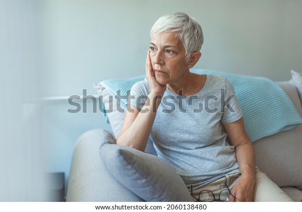 Upset depressed\
senior gray-haired lady touching temple, sitting on sofa, looking\
in the window, feeling worried and stressed about the future and\
thinking about own\
problems