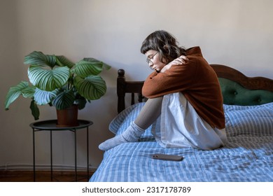 Upset depressed teen girl sitting on bed next to smartphone, feeling lonely and frustrated, sad teenager suffering from online bullying. Depression and teen use, cyberbullying concept
