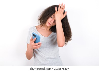 Upset Depressed Young Beautiful Brunette Teen Girl Wearing Grey T-shirt Over White Wall Makes Face Palm As Forgot About Something Important Holds Mobile Phone Expresses Sorrow And Regret Blames