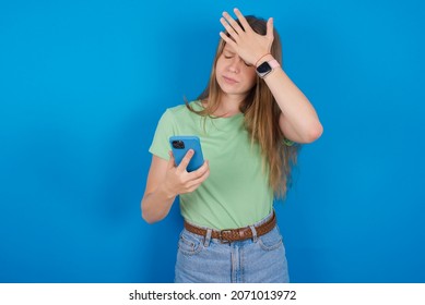 Upset Depressed Young Caucasian Girl Wearing Green T-shirt Over Blue Background Makes Face Palm As Forgot About Something Important Holds Mobile Phone Expresses Sorrow And Regret Blames