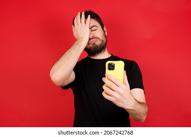 Upset Depressed Young Caucasian Man Wearing Black T-shirt Over Red Background Makes Face Palm As Forgot About Something Important Holds Mobile Phone Expresses Sorrow And Regret Blames