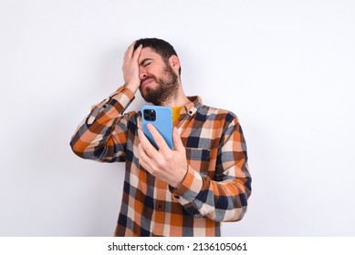 Upset Depressed Young Caucasian Man Wearing Plaid Shirt Over White Background Makes Face Palm As Forgot About Something Important Holds Mobile Phone Expresses Sorrow And Regret Blames