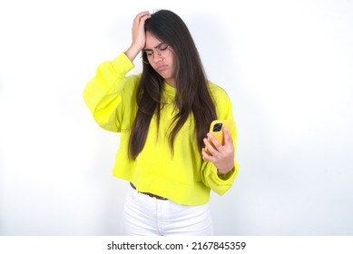 Upset Depressed Young Caucasian Woman Wearing Yellow Sweater Over White Background Makes Face Palm As Forgot About Something Important Holds Mobile Phone Expresses Sorrow And Regret Blames