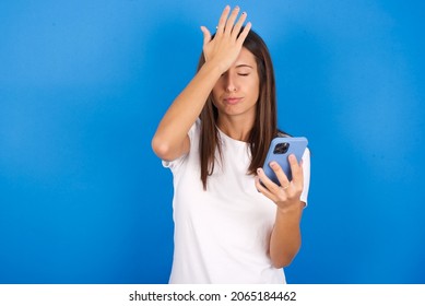 Upset Depressed Young European Woman Wearing White T-shirt On Blue Background Makes Face Palm As Forgot About Something Important Holds Mobile Phone Expresses Sorrow And Regret Blames