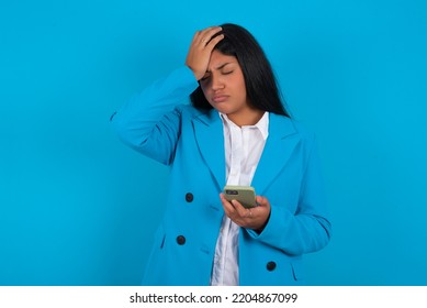 Upset Depressed Young Latin Woman Wearing Blue Blazer Over Blue Background Makes Face Palm As Forgot About Something Important Holds Mobile Phone Expresses Sorrow And Regret Blames