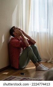 Upset Depressed Young Man Sitting With Closed Eyes On Floor At Home, Getting Unexpected Bad News Over Phone. Frustrated Guy Suffering From Relationship Break Up, Cannot Forget About Ex-girlfriend