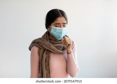 Upset despaired young indian lady in protective mask and scarf coughs, feeling bad and suffers from illness on gray background. Flu, cold, covid-19 virus and health problems. New normal, quarantine