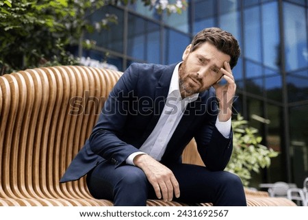 Upset and disappointed young businessman man sits on a bench near an office and court building and holds his head thoughtfully.