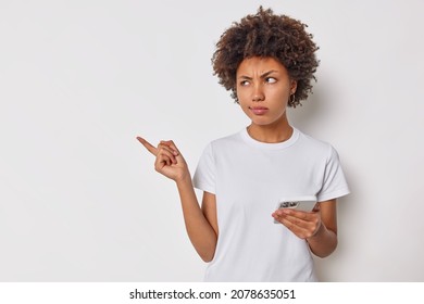 Upset discontent woman with curly hair points away with index finger shows blank copy space for your advertising uses smartphones dressed casually isolated over white background doesnt like something