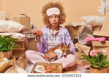Upset displeased woman feels tired after packing things in boxes prepares for relocation eats pizza poses with favorite dog surrounded by cardboard boxes with household items. Moving concept