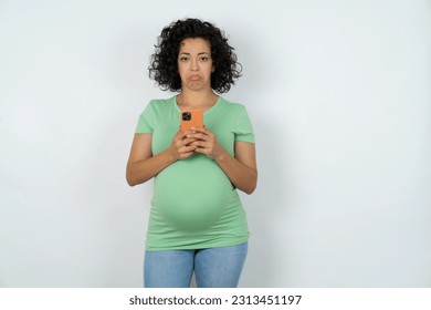 Upset dissatisfied Beautiful pregnant woman wearing green T-shirt standing over white studio background uses mobile software application and surfs information in internet, holds modern mobile hand - Powered by Shutterstock