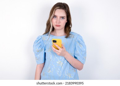 Upset dissatisfied young caucasian woman wearing blue T-shirt over white background uses mobile software application and surfs information in internet, holds modern mobile hand
