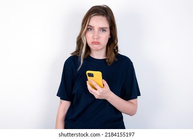 Upset dissatisfied young caucasian woman wearing black T-shirt over white background uses mobile software application and surfs information in internet, holds modern mobile hand