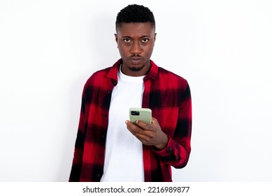 Upset dissatisfied young handsome man wearing red plaid shirt over blue background uses mobile software application and surfs information in internet, holds modern mobile hand