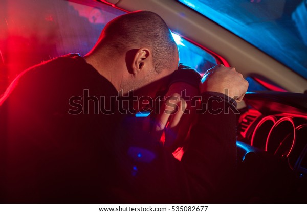 Upset drunk\
driver is caught driving under alcohol influence. Man covering his\
face from police car\
light.