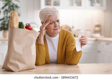 Upset Elderly Woman With Paper Bag With Food Checking Grocery Check And Touching Her Head, Sitting Alone In Kitchen, Copy Space. Old Lady Got Shocked With High Prices For Grocery