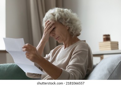 Upset Elderly Woman Sitting On Couch At Home, Reading Document And Getting Stressful Bad News At Home. Frustrated Senior Lady Shocked With Letter, Touching Head, Trying To Cope With Negative Emotions