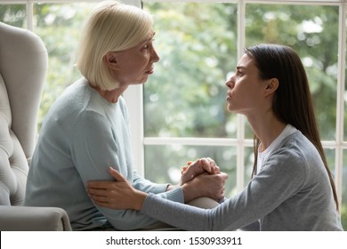 Upset Empathic Young Lady Sitting Near Chair, Holding Hand Of Desperate Unhappy Middle Aged Mommy, Showing Support And Care. Side View Grown Up Daughter Giving Psychological Help To Stressed Mom.