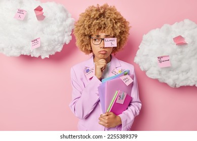 Upset female corporate worker keeps hand on chin looks unhappily aside wears transparent eyeglasses and formal clothing poses with folders uses stickers for writing down list to do. Office work - Shutterstock ID 2255389379