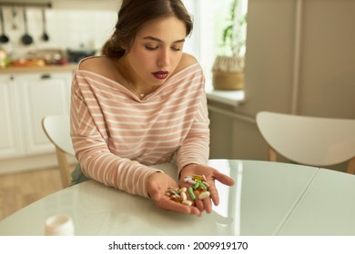 Upset female looking at handful of drugs with sadness. Young sad woman sitting at table in home kitchen. Weight loss pills and dietary supplements concept - Shutterstock ID 2009919170
