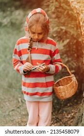 Upset Or Focused Eight Year Old Kid Girl Mushroom Picker Is Seek For And Picking Mushrooms In The Forest. Child Survivalists Gathers A Wild Fungus Harvest And Outdoor Foraging  In Nature. Flare