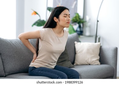 Upset frustrated asian young woman suffering from backache, unhappy japanese brunette sitting on a sofa in living room, feeling discomfort because of pain in back, need treatment and rest