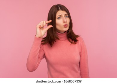 Upset frustrated woman with brown hair in pink sweater showing small size with her fingers, sceptic about centimeter inch size, measuring scale. Indoor studio shot isolated on pink background