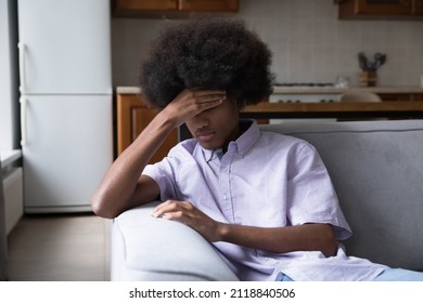 Upset Fuzzy Haired Black Teen Guy Sitting On Couch, Touching Head, Feeling Bad, Frustrated, Sad, Depressed, Suffering From Head Ache, Going Through Stress, Emotional Crisis, Trauma