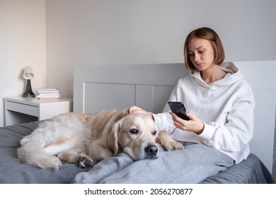 An Upset Girl Sits With A Dog On The Bed, Looks At The Phone And Listens To Sad Music. Teenage Depression.