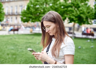 An upset girl types a message on her phone. The girl writes a text message on the street against the background of green trees. - Shutterstock ID 2160162723