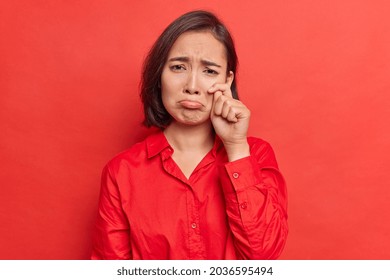 Upset gloomy Asian woman wipes tears after watching sad touching movie has heartbroken feelings feels discontent wears shirt poses against bright red background. Female model feels down depressed