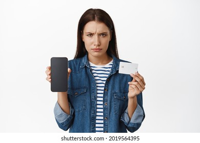 Upset Gloomy Woman Showing Mobile Phone Screen, Empty Display On Smartphone And Credit Card, Furrow Eyebrows Disappointed, Zero Money On Bank Account App, White Background