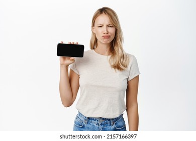 Upset And Gloomy Young Woman Showing Horizontal Mobile Phone Screen, Grimacing Disappointed, Complaining At Smth, Standing Over White Background