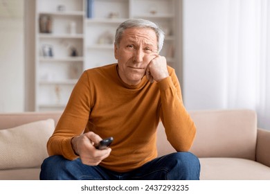 Upset grey-haired european elderly man with remote control in his hand sitting on couch at home, switching channels on TV, feeling bored, nothing to watch, copy space - Powered by Shutterstock