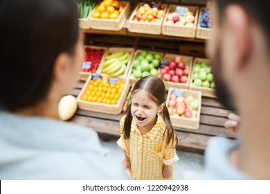 Upset hysterical girl with closed eyes crying loudly while manipulating parents and standing against food stall in supermarket