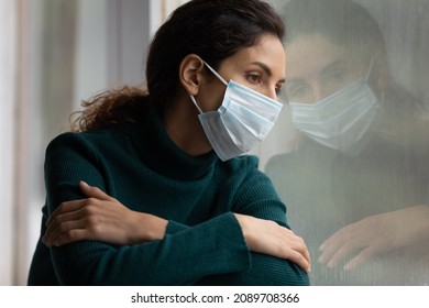 Upset Ill Hispanic Woman In Facemask Look In Window Distance Feel Sad Lonely On Lockdown At Home. Sick Sad Latino Female In Facial Mask Suffer From Loneliness Solitude On Covid-19 Quarantine.