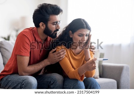 Upset indian couple with negative pregnancy test sitting on couch at home, hugging and looking at result, depressed eastern man and woman suffering from infertility problem, copy space